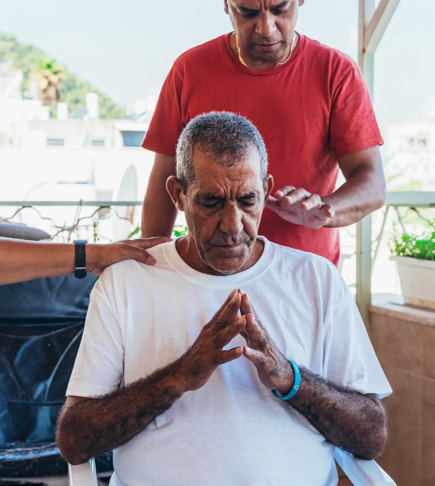 A man in a red shirt lays hands on and prays for another man who is sitting in a chair on a balcony.